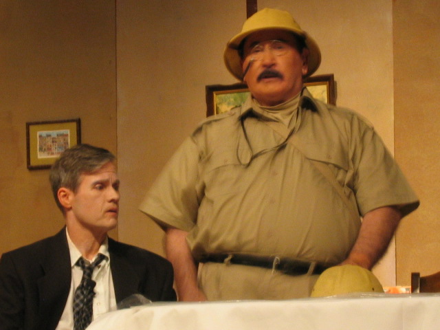 Man in pith helmet standing next to seated suited man.