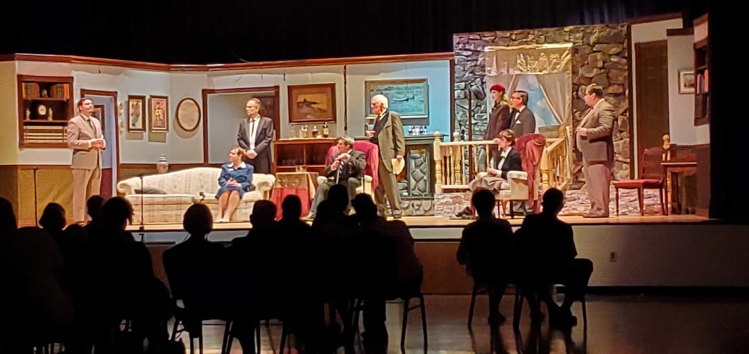 A man in a light brown suit, a woman in a blue skirt sitting on a couch, a man in a light gray suit standing behind her, a man
       in a dark gray suit sitting in a chair, a man in a grayish-green suit standing behind him, a woman in a red cap, a woman in a dark
        jacket sitting in a chair, a man in a dark gray suit, and a man in a brown suit