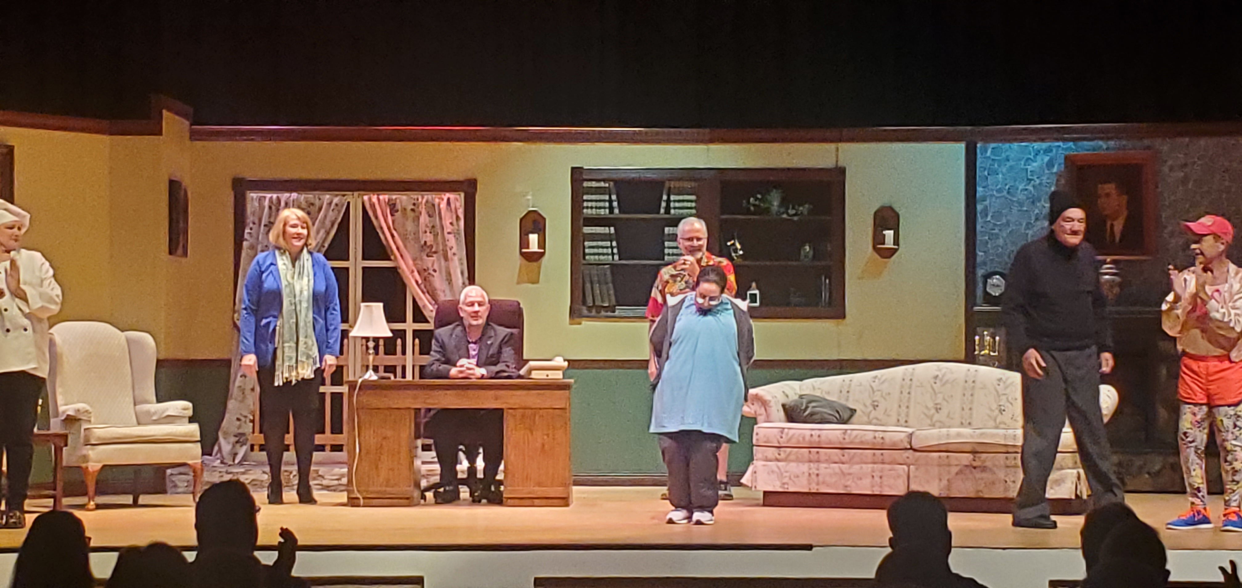 From left to right: a chef in a tope, a woman in a blue jacket and light green scarf, a man in a black suit sitting behind a desk, a woman in a long bluish green top standing in front of a man in a red Hawaiian shirt and brown shorts,
       a man in a black pullover, gray pants and black cap, and a man in an orange running outfit clapping.