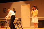 A man in a white shirt with his back to her, talking to a cringing woman in a yellow sweater