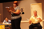 A standing woman reading from a script, in front of a sitting man with a script, with another man at a table