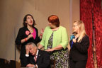 A man sitting at a small desk and three women standing, two in black pantsuits one in a green sweater