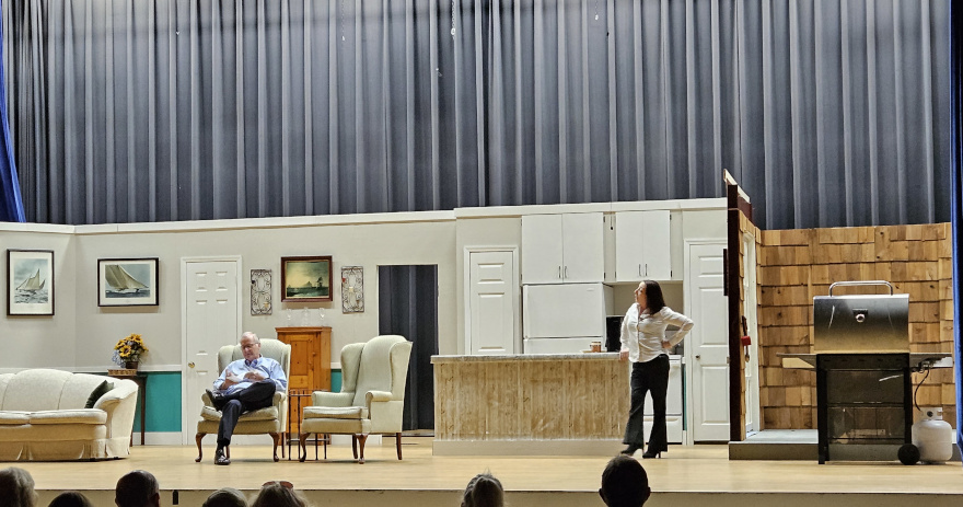 A man in a blue shirt sitting in a chair reading and a woman in a white blouse and black pants standing.