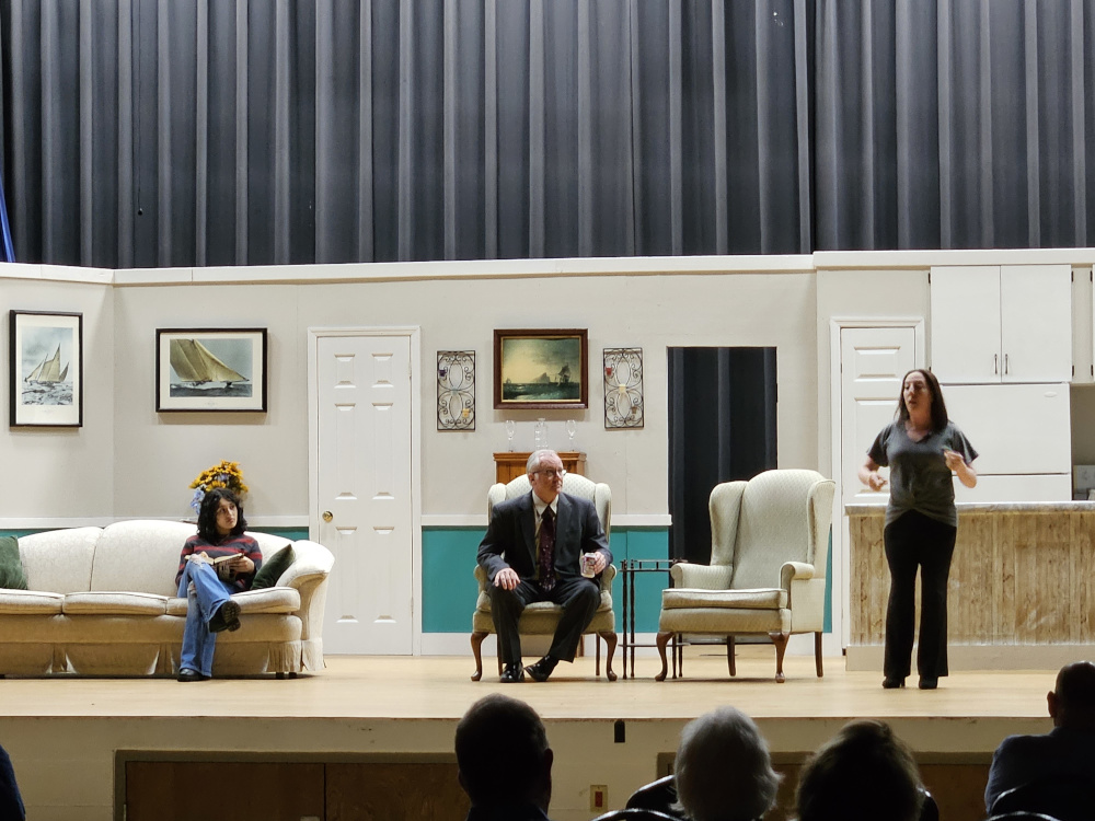 A girl sitting on the couch reading, a man sitting in a chair wearing a dark gray suit, and a woman standing in front of the kitchen.