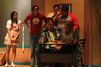 A man and a woman sit at a desk, while a young woman, a man in a red Avengers T-shirt, a man in a suit,
        and a man in a Tampa Bay Buccaneers T-shirt look on behind them