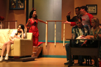 A young woman sits on a couch, while a woman dressed like a devil talks to a man in a suit and a man in a Tampa Bay Buccaneers T-shirt, and
       a man a woman sit at a desk