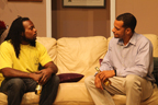 Two men on a couch facing one another, one wearing a yellow T-shirt, one holding a beer