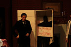 A man in a dark suit standing fron front of a tall box with a sign that says The Doom of Devilsmoor