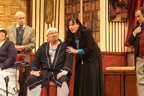 A woman in a dark dress and blue blouse stands behind a bald man with a bandage on his head, while
       Sherlock Holmes looks on from the right, and Dr. Watson and a woman in gray vertical striped dress look on from the left