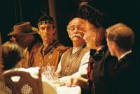 A man in a cowboy hat, a Native American man, a bald man, a woman in a hat, a man in a suit, and a woman with her back
       to the camera all sitting a table