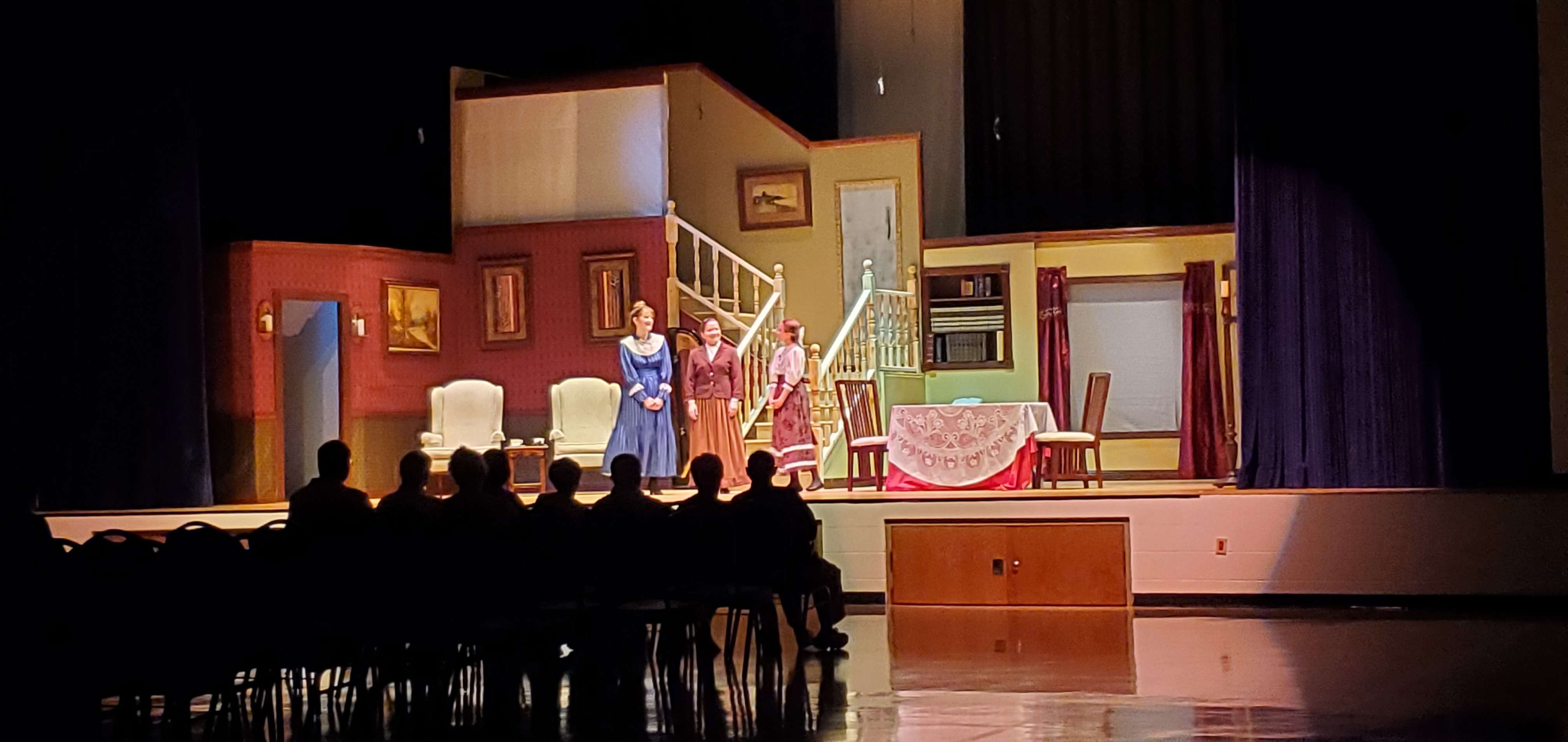 A very wide shot of a woman in a long blue dress to the left, a woman in a reddish-brown jacket and a light brown dress in the middle and a girl in a brown dress to the right.