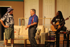 A man in a cloth hat talking to a man in red suspenders while another man with a guitar looks on