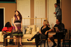 A man in a Bob marley shirt on the couch, a woman standing, another woman sitting, a man sitting reading a book, and another man stepping into the room from
       an open door