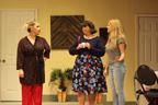 A woman in a red polka dot with dark background robe and red pants, a woman in a dark blouse and floral dress talks to a young woman in a gray T-shirt and jeans