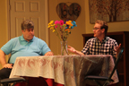 A man in a blue shirt talking to a young man in glasses and a plaid shirt at a table with a vase of flowers between them