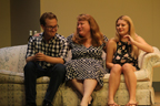 A young man in glasses and a plaid shirt, a woman in a gray and black dress and a young woman in a dark floral print dress all sitting on a couch